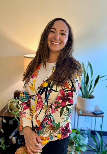Basak Kaya smiling, wearing a colorful floral jacket, in a bright room with houseplants